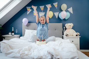 Happy emotional woman in pajama and party cap after blowing out the candles on birthday cake, sitting on the bed in decorated bedroom. Make a wish process. Morning surprise. Happy birthday concept.