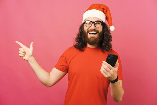 Young Christmas man points aside while holding a phone and wide smiling.