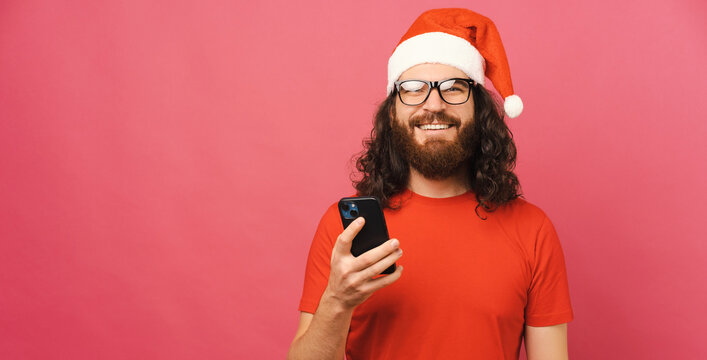Long haired bearded man with Christmas hat is holding a phone and smiles.