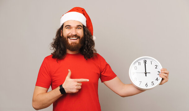 Young man is pointing at a round white clock while wearing a Christmas hat.