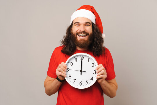 Young bearded man is holding a round white clock while wearing a Christmas hat.