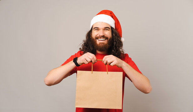 Cheerful young bearded man wearing Christmas hat is holding a paper bag.
