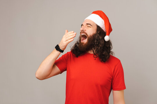 Young bearded man yawns while wearing a Christmas cap over grey background.