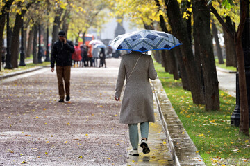 Rain in autumn season, woman with umbrella walking on falling leaves in city park on people background