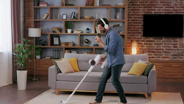 Cheerful guy, the man cleans the apartment. Vacuuming, wearing headphones with dancing and singing into a smartphone instead of a microphone. House cleaning is easy.