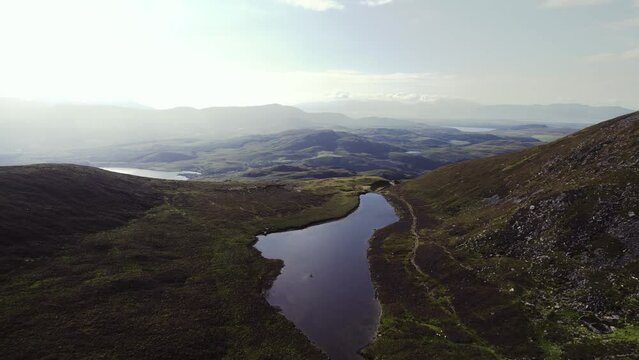 Beautiful aerial of irish highlands with a lough laying within grass covered mountains. Revealing grassy mountains and valleys are behind in the distance. Flying fowards during evening hour in summer.