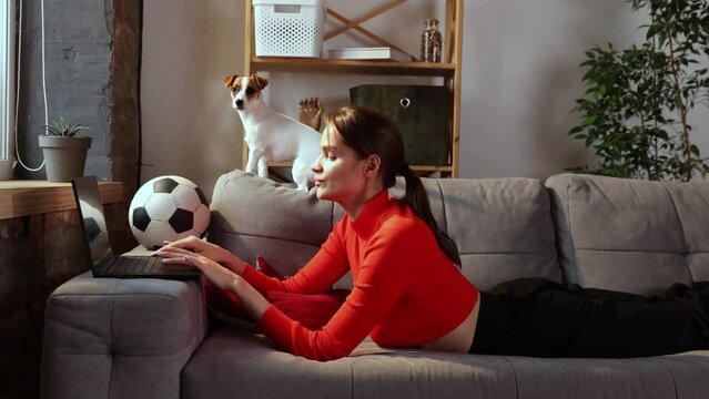 Young woman is using laptop and lying with Jack Russell Terrier dog at home. Beautiful girl hugging with her pet and looking at computer screen with smile. Domestic animal, football fan concept