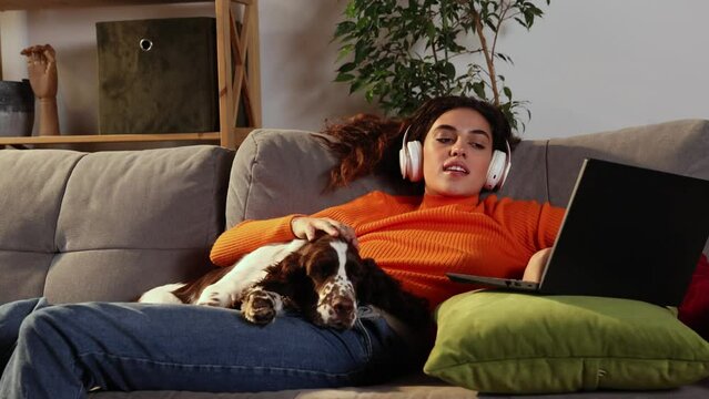 Young woman is using laptop and lying with English springer spaniel dog on bed at home. Beautiful girl hugging with her beloved pet and looking at computer screen with smile. Domestic animal