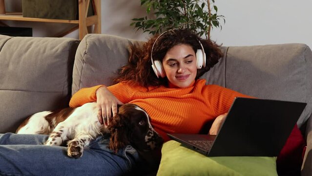 Young woman is using laptop and lying with English springer spaniel dog on bed at home. Beautiful girl hugging with her beloved pet and looking at computer screen with smile. Domestic animal