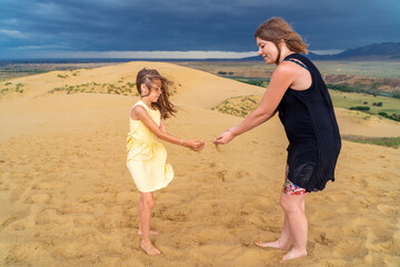 Happy girl in yellow dress with mother stands with sand falling through their hands at sandy dune...