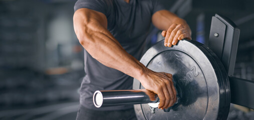 Bodybuilder Putting Barbell Plates on the Bar. Focus on barbell plate, close up shot male strong hands adding weight to barbell at gym. Wide background banner for gym, fitness workout, bodybuilding