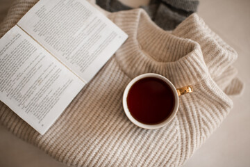 Cup of coffee stay on stack of knitted textile sweater in bed with paper open book close up. Winter cozy season. Top view.