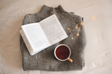 Cup of coffee stay on knitted textile sweater in bed with paper open book with christmas glowing...