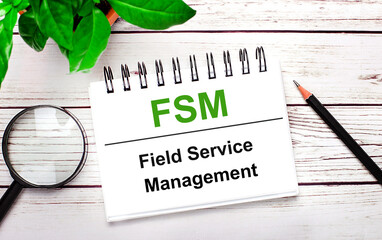 On a light wooden background, a magnifying glass, a pencil, a green plant and a white notebook with text FSM Field Service Management. Business concept