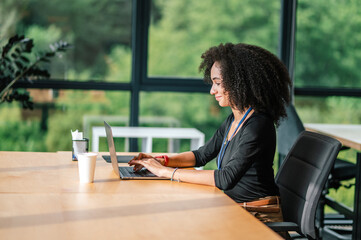 Curly-haired young woman sitting at the table in the office and working on a laptop