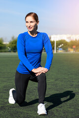 Women and sport. Girl in sportswear does exercises: kneels and stretches on the grass at the stadium on a sunny day. Middle aged sportswoman dressed in sportsclothes exercising outdoor