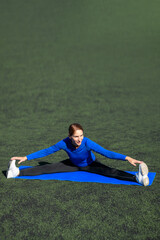 Women and sport. Smiling Girl in sportswear - blue shirt and black leggings does exercises: do twine on the grass at the stadium outdoor on a sunny day. Middle aged sportswoman 