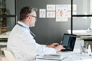 Fototapeta na wymiar Mature medical professional examining x-ray image on laptop at table at his office