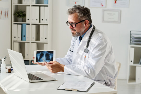 Mature doctor in white coat showing x-ray image to patient and discussing treatment online using laptop at table at office