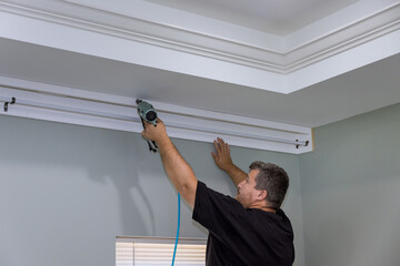 There is professional handyman who is nailing wooden cornices on the curtain wall over window of a newly constructed home © ungvar
