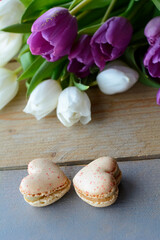 Fototapeta na wymiar Two tasty creamy macarons are lying on the wooden table with white and violet tulips. Two macarons in form of the heart as 8. Dessert is on the wood