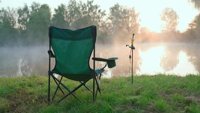 River at dawn with foggy haze with fishing gear on the shore. Camping chair and spinning for fishing. Beautiful sunrise - morning landscape. Place of a fisherman with fishing rod on lake into the fog