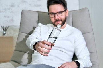 Portrait of handsome bearded man in glasses, young thirsty person is drinking pure water from plastic bottle at home sitting at comfortable armchair, show bottle 