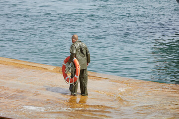 sailor in the port with an orange lifebuoy in his hands. He is dressed in a green waterproof suit.
