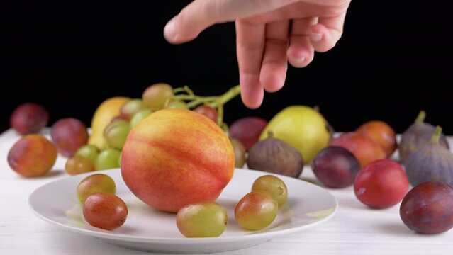 Female Hand Putting a Ripe Apple or Peach on a Plate with Grape and Fruit. Many different colorful fruits, plums, pears lie on the table. Harvest. Black and white background. Variety exotic products.