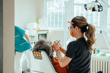 Obraz premium Close up photo of woman dentist in protective workwear using a dental drill for treating dental cavity on male patient teeth in dental clinic with dental assistant in the background