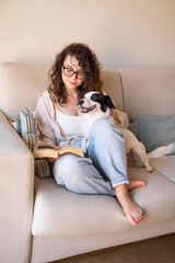 Obraz na płótnie Canvas Beautiful curly girl with glasses sitting on sofa with book and her black and white adopted dog. Light sunny living room. reading with smiling pet. Happy relaxed weekend moment at home with dog