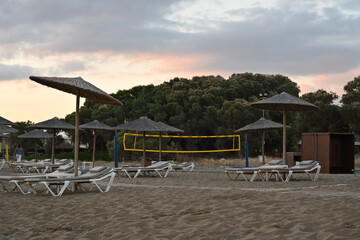 Fototapeta premium Sandy beach of a holiday resort with plastic deck chairs and straw beach umbrellas during sunrise. Behind is playground for beach volleyball with yellow net under clouds.