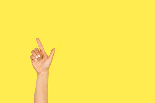 Mano de mujer con el brazo y la mano levantada apuntado con el dedo indice hacia arriba sobre un fondo amarillo liso y aislado. Vista de frente y de cerca. Copy space