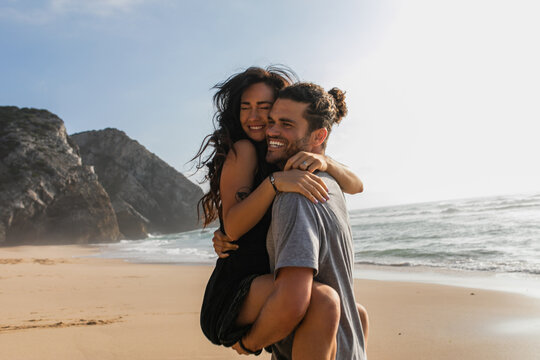 cheerful man lifting and hugging tattooed woman in dress on beach near ocean.