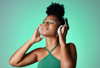 Black woman in green studio with headphones on, listening to music. Young happy girl with makeup,...