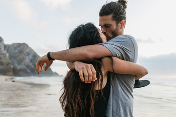 bearded man and brunette woman hugging and kissing on beach in portugal.