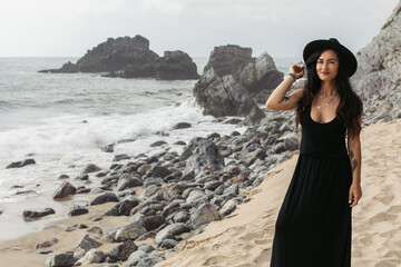 cheerful and tattooed woman in black dress and hat standing on sandy beach in portugal.