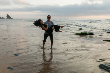 happy man lifting cheerful girlfriend on dress while standing on ocean water.