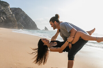 bearded man smiling while lifting woman in dress and having fun on beach near ocean. - Powered by Adobe