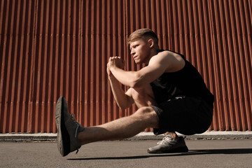 Horizontal full body shot of strong Caucasian man wearing black outfit doing one leg bodyweight squat exercise outdoors