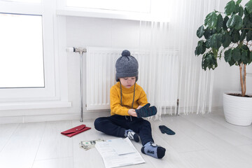 A little boy in a yellow sweater and hat is counting money and studying heating bills, near a heater with a thermostat.