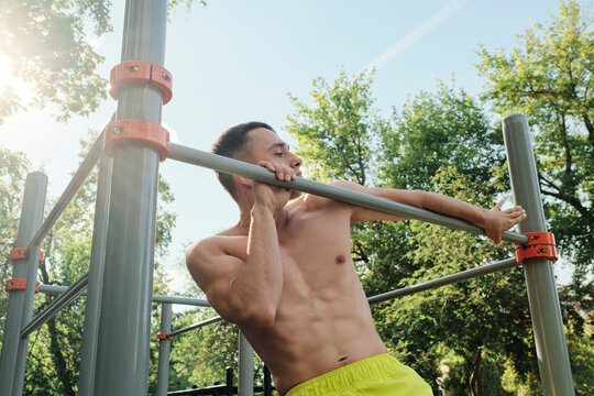 Portrait of handsome shirtless young man exercising in park on pull-up and dip bar on summer day