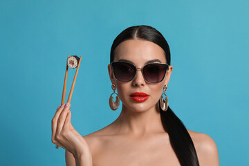 Attractive woman in fashionable sunglasses holding chopsticks with sushi against light blue...