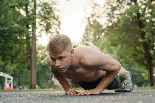 Horizontal shot of shirtless young man with strong body spending summer morning in local city park doing strengthening workout, practicing diamond plank