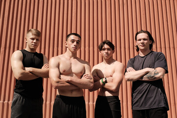 Horizontal medium portrait of four sporty men with muscular bodies wearing comfortable clothes standing with arms crossed looking at camera