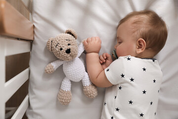 Adorable little baby with pacifier and toy sleeping in crib, top view