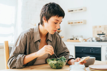 A young brunette is sitting in the kitchen eating and browsing social networks on her phone.