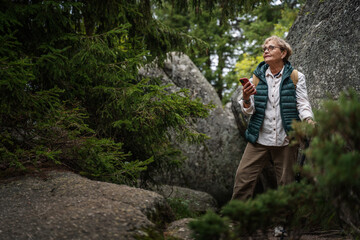 Naklejka na ściany i meble Happy active senior mature woman walking in the forest and using smartphone for navigation. Travel, active retirement and healthy lifestyle concept