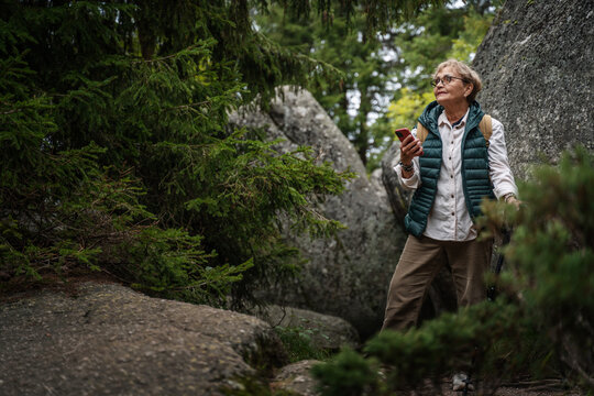 Happy active senior mature woman walking in the forest and using smartphone for navigation. Travel, active retirement and healthy lifestyle concept