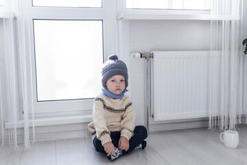 A small child in a sweater and a gray hat is kneeling near a heater with a thermostat.